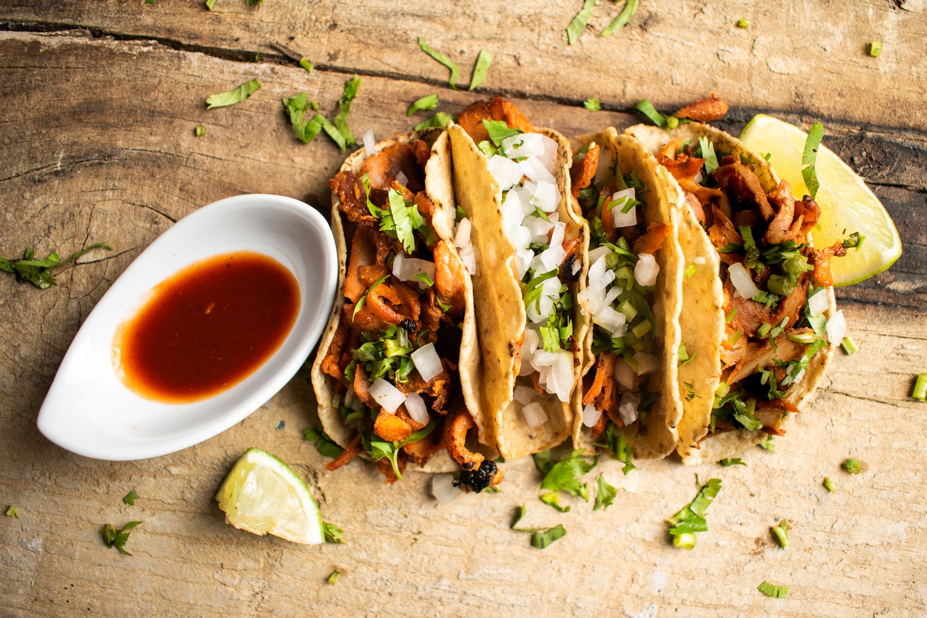 A row of al pastor tacos on corn tortillas with onion, cilantro and lime, served with a hot sauce.